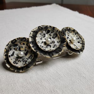 May include: A decorative brooch featuring three fabric flower designs with a leopard print pattern in black and white. Each flower has a faux pearl center. The brooch is attached to a silver-tone pin.