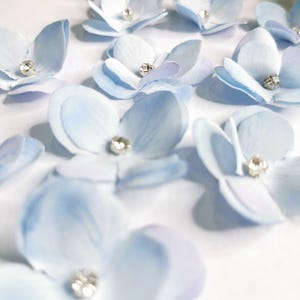 Könnte beinhalten: Hellblaue Stoffblumen mit einem funkelnden Strassstein in der Mitte. Die zarten Blütenblätter sind auf einer weißen Oberfläche angeordnet, mit grünem Laub im Hintergrund. Diese floralen Verzierungen eignen sich für Bastelarbeiten und Dekorationen.