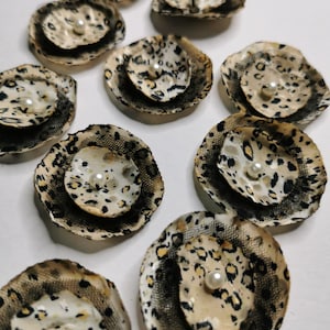 May include: A collection of handmade fabric flowers with a leopard print pattern. Each flower has a central pearl and is trimmed with black lace. The flowers are arranged on a white surface.