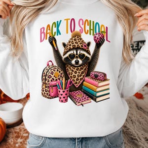 Peut inclure: Un sweat-shirt blanc avec une illustration de raton laveur portant un bonnet, une écharpe et des lunettes en forme de cœur à motif léopard. Le raton laveur tient un crayon et une pomme. On voit un sac à dos, un pot à crayons et des livres. Texte : BACK TO SCHOOL.
