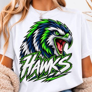 Puede incluir: Una camiseta blanca de manga corta con un gran gráfico de una cabeza de halcón con plumas azules, verdes y blancas. El halcón tiene el pico abierto y ojos penetrantes. Debajo del ave, la palabra HAWKS está impresa en una fuente blanca dentada.