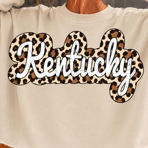 May include: A beige long-sleeve sweatshirt with a raw-edge hem. The front features the word Kentucky in white script lettering, layered over a brown and black leopard print graphic shape with a rounded, bubbly outline design.
