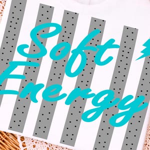 May include: White t-shirt with a design featuring gray vertical stripes dotted with black circles. The words "Soft Energy" are written in a teal script font across the center of the shirt. The shirt is displayed on a woven basket.