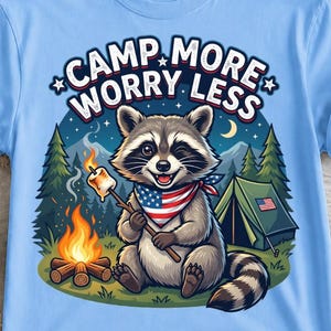 Peut inclure: Un t-shirt bleu clair avec une illustration de raton laveur portant un bandana aux couleurs du drapeau américain, grillant une guimauve sur un feu de camp. Le texte CAMP MORE WORRY LESS est inscrit au-dessus, avec une tente et des sapins.