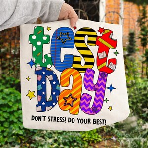 Peut inclure: Un sac fourre-tout crème avec les mots "TEST DAY" en lettres colorées et à motifs. Le sac présente des étoiles et le texte "DON'T STRESS! DO YOUR BEST!" en noir. Le sac est tenu par une personne.