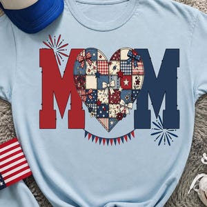 Peut inclure: Un t-shirt bleu clair avec le mot MOM en grandes lettres majuscules, où un cœur en patchwork remplace la lettre O. Le motif du cœur présente des carreaux, des étoiles et des nœuds rouges, blancs et bleus. Des feux d'artifice et des guirlandes.