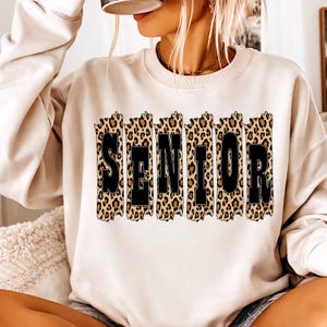 Peut inclure: Sweat-shirt beige avec le mot "SENIOR" en lettres noires. Les lettres sont placées sur un fond à imprimé léopard. Le sweat-shirt est de couleur neutre et a un col rond.