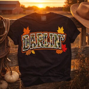 Peut inclure: T-shirt noir avec le mot "DARLIN'" en motif camouflage, bordé de blanc et d'orange. Le design comprend des feuilles d'automne orange, jaune et rouge. Un t-shirt classique à col rond.