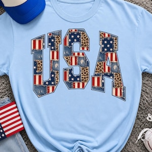 Peut inclure: Un t-shirt bleu clair arborant les lettres USA dans une grande police de caractères. Les lettres sont remplies d'un motif patchwork composé de rayures du drapeau américain, de textures en jean bleu et d'un imprimé léopard brun et noir.