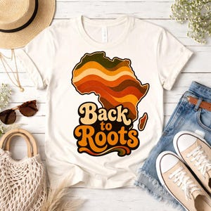 Peut inclure: T-shirt crème avec un motif rétro du continent africain en marron, orange et vert. Le texte "Back to Roots" est en dessous. Le t-shirt est associé à un short en jean et des baskets en toile.
