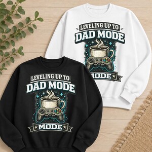 Peut inclure: Un sweat-shirt noir et un sweat-shirt blanc, chacun arborant une impression graphique d'une tasse de café posée sur une manette de jeu vidéo avec le texte "LEVELING UP TO DAD MODE" et "MODE" dans une police stylisée avec des étoiles et des accents bleus.