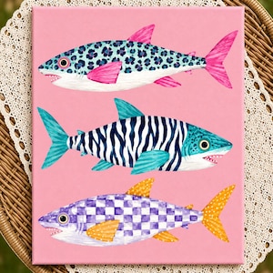 Peut inclure: Une impression sur toile rectangulaire représentant trois requins sur un fond rose uni. Le requin du haut a un motif léopard bleu avec des nageoires roses. Le requin du milieu a des rayures zébrées bleues et blanches. Le requin du bas a un damier violet.