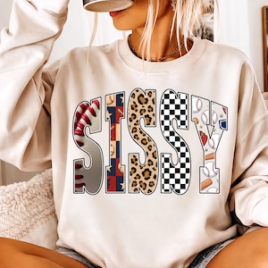 Peut inclure: Sweat-shirt beige avec le mot "SISSY" dans un design audacieux et à motifs multiples. Les lettres présentent des coutures de baseball, un imprimé léopard, un motif en damier et des graphiques sur le thème du baseball. Le sweat-shirt a un col rond.