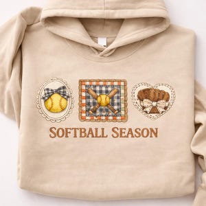 Puede incluir: Sudadera con capucha beige con la inscripción marrón "SOFTBALL SEASON". El diseño incluye tres parches bordados: una pelota de softbol con un lazo, un bate y una pelota de béisbol, y un guante de softbol con un lazo. Los parches están enmarcados con bordes decorativos.