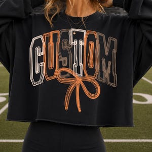 Peut inclure: Un sweat-shirt noir court avec le mot CUSTOM écrit dans une police de style gribouillé superposée en blanc, orange et marron. Sous le texte se trouve un grand motif de nœud orange dessiné à la main. Le sweat-shirt a un ourlet brut.