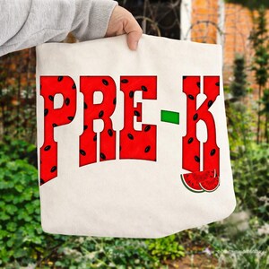 Peut inclure: Un sac fourre-tout en toile couleur crème avec le texte PRE-K en grandes lettres grasses. Les lettres sont remplies d'un motif de pastèque rouge avec des pépins noirs. Un petit tiret vert sépare les lettres et une tranche de pastèque est en bas.