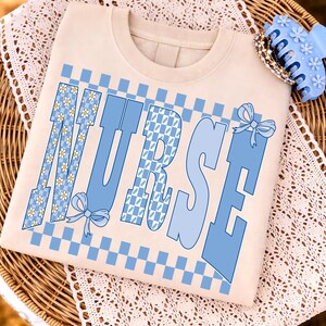 Peut inclure: Un sweat-shirt beige à col rond avec le mot "NURSE" en grandes lettres bleues. Les lettres ont un motif à carreaux et des accents floraux. Une pince à cheveux bleue et un bracelet sont sur le côté.