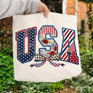 Peut inclure: Sac fourre-tout crème avec le mot "USA" dans un design patriotique. Les lettres sont remplies de motifs, notamment des étoiles, des rayures, des imprimés floraux et un nœud vichy. Les bords des lettres ont une bordure festonnée.