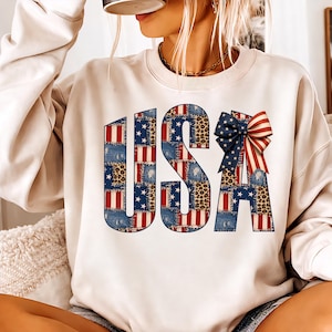 Peut inclure: Un sweat-shirt crème avec le mot "USA" dans un motif patchwork. Les lettres incorporent des motifs de rayures rouges et blanches, d'étoiles bleues, de denim et d'imprimé léopard. Un nœud patriotique est attaché à la lettre "A".