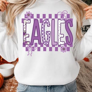 May include: A white long-sleeve sweatshirt featuring the word EAGLES in large purple letters. The design includes a purple and white checkered background, floral patterns on the letter E, a plaid pattern on the letter A, and small bows and flowers.