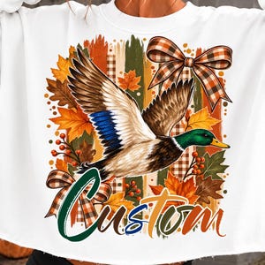 Peut inclure: Un sweat-shirt blanc à manches longues avec une impression graphique représentant un canard colvert en vol, entouré de feuilles d'automne, de baies et de nœuds à carreaux. Le mot "Custom" est écrit en police script multicolore.