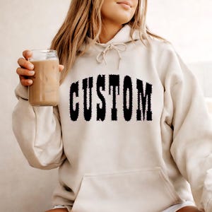 Peut inclure: Sweat à capuche crème avec le mot "CUSTOM" en noir, souligné d'un motif texturé. Le sweat à capuche a une capuche à cordon et une poche avant. La personne tient un verre de café glacé.