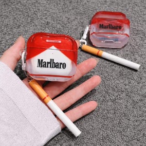 Puede incluir: Fundas para AirPod rojas y blancas de la marca Marlboro con un llavero con forma de cigarrillo. Las fundas son transparentes y tienen una parte superior roja con el logotipo de Marlboro. Los llaveros de cigarrillos son blancos con un filtro naranja.