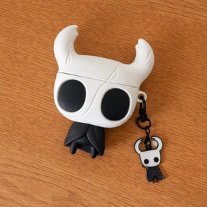 May include: A white and black earbud case with a cartoon character design, including large eyes and horns. It has a matching charm on a black clip. The case features a black cape-like design at the bottom.