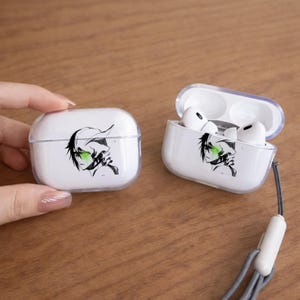 May include: White AirPods Pro cases with a clear lid, featuring a black and green anime character design. One case is open, revealing white earbuds. A grey lanyard is attached to the open case. The cases are on a wooden surface.