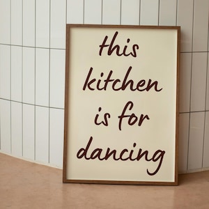 Könnte beinhalten: Ein gerahmtes Schild mit den Worten "this kitchen is for dancing" in dunkelroter Schrift. Das Schild hat einen hellcremefarbenen Hintergrund und einen braunen Holzrahmen. Der Hintergrund zeigt weiße rechteckige Fliesen.