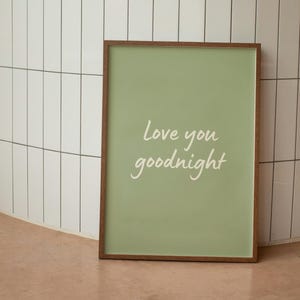 以下が含まれることがあります： 明るい緑色の背景に「Love you goodnight」の文字が白で書かれた、額入りのアートプリント。 フレームは温かみのある茶色です。 プリントは白いタイルの壁に立てかけられています。