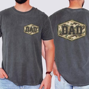 May include: Charcoal grey t-shirt featuring the word "DAD" in a camouflage design, displayed on the front and back. The text is within a hexagonal frame. The shirt has a crew neck and short sleeves.