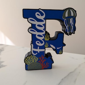 Op de afbeelding: Een blauwe houten letter "F" met de naam "Fedde" in witte letters. De letter is versierd met een onderwaterthema, inclusief een kwal, koraal en zeewier.