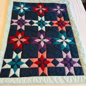 Puede incluir: Una colcha rectangular con un fondo azul marino oscuro y un patrón repetitivo de estrellas de ocho puntas multicolores en tonos rojo, rosa, morado y verde azulado. La colcha está rematada con un borde de tela color verde menta claro.