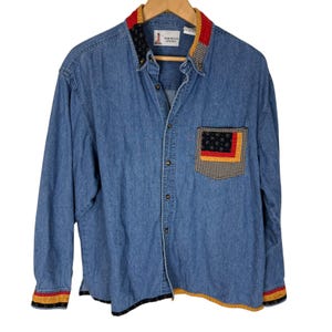 May include: A blue denim shirt with long sleeves and a collared neckline. The shirt has a pocket with a patterned design and decorative trim on the collar and cuffs. The shirt is hanging on a wooden hanger.