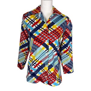 Op de afbeelding: Een overhemd met knopen met een levendig geometrisch patroon in rood, geel, blauw en wit. Het shirt met lange mouwen heeft een kraag en een mix van ruitjes, pied-de-poule en stippen. Het shirt wordt gepresenteerd op een witte mannequin.