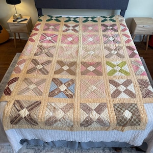 Op de afbeelding: Een patchwork quilt met een raster van vierkante blokken, elk met een wit vierkant in het midden omringd door vier rechthoekige stukken in diverse patronen en kleuren, waaronder roze, bruin, blauw, groen en crème, omlijst door lichtbruine banen.