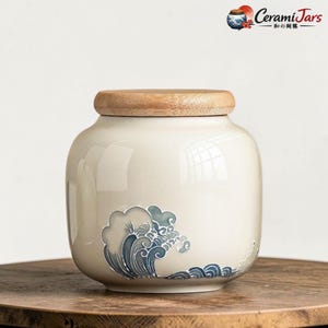 Może przedstawiać: Kremowy ceramiczny słoik z drewnianą pokrywką i niebieskim wzorem fal. Słoik stoi na drewnianej powierzchni. Logo "CeramiJars" znajduje się w prawym górnym rogu.