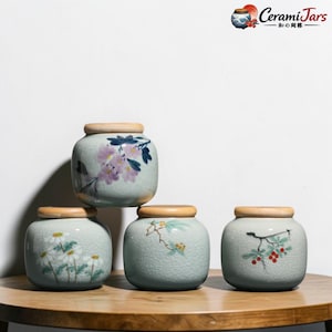 Puede incluir: Cuatro tarros de cerámica con tapas de madera se exhiben sobre una superficie de madera. Los tarros son de color azul claro con diseños florales en rosa, blanco y verde. Los tarros son cuadrados con bordes redondeados.