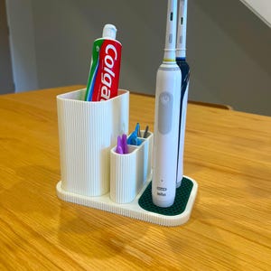 May include: A white toothbrush holder with a ribbed design, holding a tube of Colgate toothpaste, two electric toothbrushes, and small grooming tools. The holder sits on a green mat.