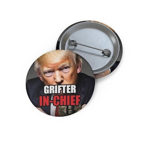 May include: A circular pin-back button featuring a portrait of Donald Trump wearing a dark suit and red tie. The text GRIFTER is printed in white and IN-CHIEF is printed in red over the image. The back shows a silver metal pin mechanism.