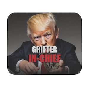 May include: A rectangular mousepad featuring a portrait of Donald Trump wearing a dark suit and red tie. The text GRIFTER is printed in white above the text IN-CHIEF in red. A gold Bitcoin coin is held in the foreground of the image.
