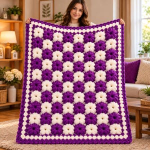 May include: A rectangular crochet blanket featuring a repeating checkerboard pattern of textured, puff-stitch flower motifs in alternating deep purple and creamy white yarn, finished with a matching purple crocheted border.