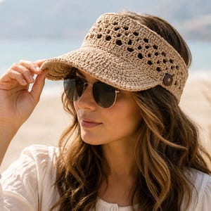 May include: A beige crochet visor featuring an openwork geometric pattern on the crown and a solid brim. The visor is accented with a small, dark brown circular button on the side, providing a secure fit while maintaining a textured, woven appearance.