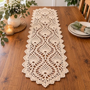 May include: A long, rectangular cream-colored crochet table runner featuring an intricate, repeating pineapple-inspired lace pattern with scalloped edges, laid flat along the center of a wooden table surface.