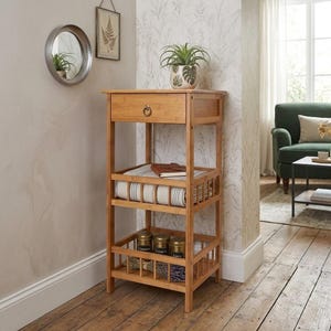 May include: A light brown bamboo storage shelf with a drawer and three open shelves. The top shelf holds a potted plant. The middle shelf has a rolled blanket and a book. The bottom shelf has jars and dried herbs.