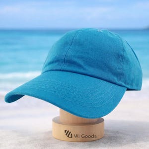 May include: A solid turquoise baseball cap with a curved brim. The cap is displayed on a wooden stand with the text "Wi Goods". The background features a beach and ocean.