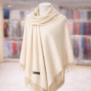 May include: A cream-colored shawl draped over a mannequin. The shawl has a soft, flowing texture with a fringed edge. A small label is visible near the bottom. The background is blurred, suggesting a retail setting. This elegant accessory is perfect for adding warmth and style.