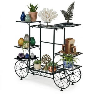 May include: Black metal plant stand with multiple shelves and decorative wheels. The stand displays various potted plants, vases, and decorative objects, including a coral sculpture, a fern statue, and bookends. The stand is designed for indoor or outdoor use.