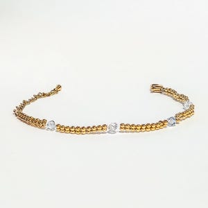 May include: A gold-colored bracelet with a delicate design. The bracelet features small, round beads and clear, square-cut crystals. The bracelet is displayed on a white background, highlighting its elegant simplicity. A piece of jewelry.